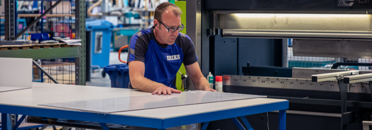 Een ERIKS-medewerker in een blauw uniform positioneert zorgvuldig een plastic plaat op een werktafel in een werkplaats, met industriële machines en stellingen op de achtergrond.