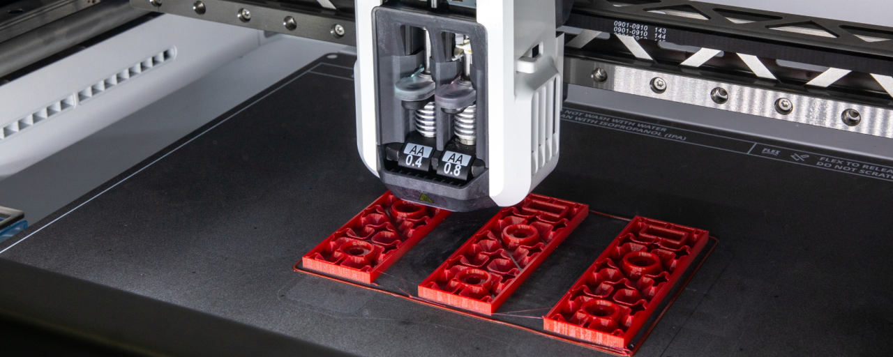 Linkedin Post - Shoes (600 x 600 px) - 1 Een close-up van een 3D-printer die rode plastic onderdelen creëert, met precieze nozzles die materiaal op het printbed aanbrengen.