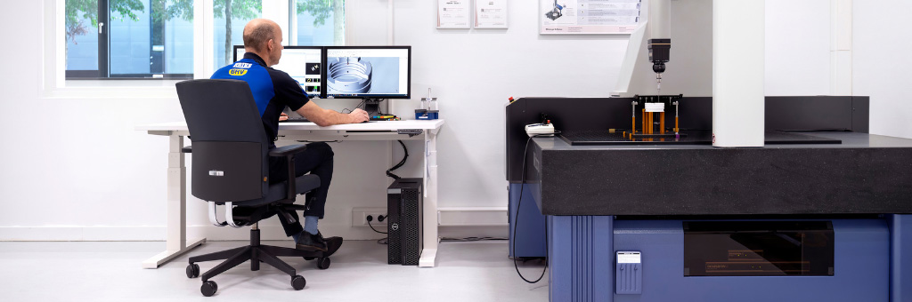 Ein ERIKS Mitarbeiter sitzt an einem Schreibtisch und arbeitet an einem Computer mit technischen Entwürfen auf dem Bildschirm, neben einer großen Präzisionsmessmaschine in einer sauberen, weißen Laborumgebung.