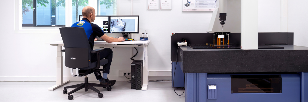 Ein ERIKS Mitarbeiter sitzt an einem Schreibtisch und arbeitet an einem Computer mit technischen Entwürfen auf dem Bildschirm, neben einer großen Präzisionsmessmaschine in einer sauberen, weißen Laborumgebung.