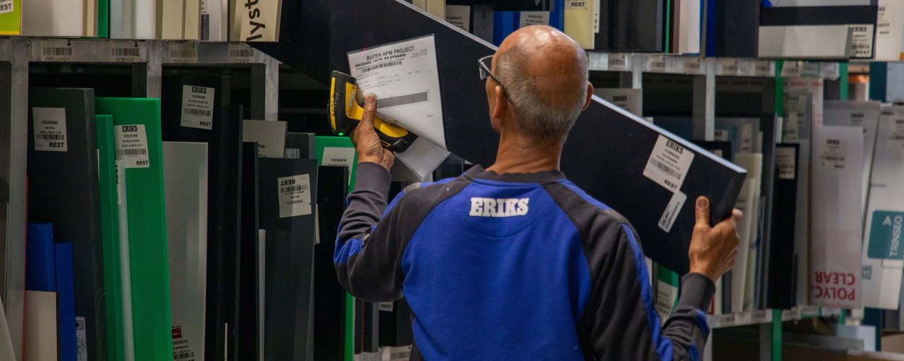 Ein ERIKS Mitarbeiter in blauer Arbeitskleidung entnimmt eine beschriftete Plastikfolie aus einem Regal, das von mehreren Mustern in verschiedenen Farben und Größen umgeben ist.
