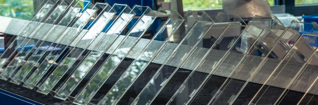 Eine Reihe von transparenten Kunststoffplatten, die auf einem Metallgestell aufrecht stehen und deren Oberflächen das Licht in einer Werkstattumgebung reflektieren.