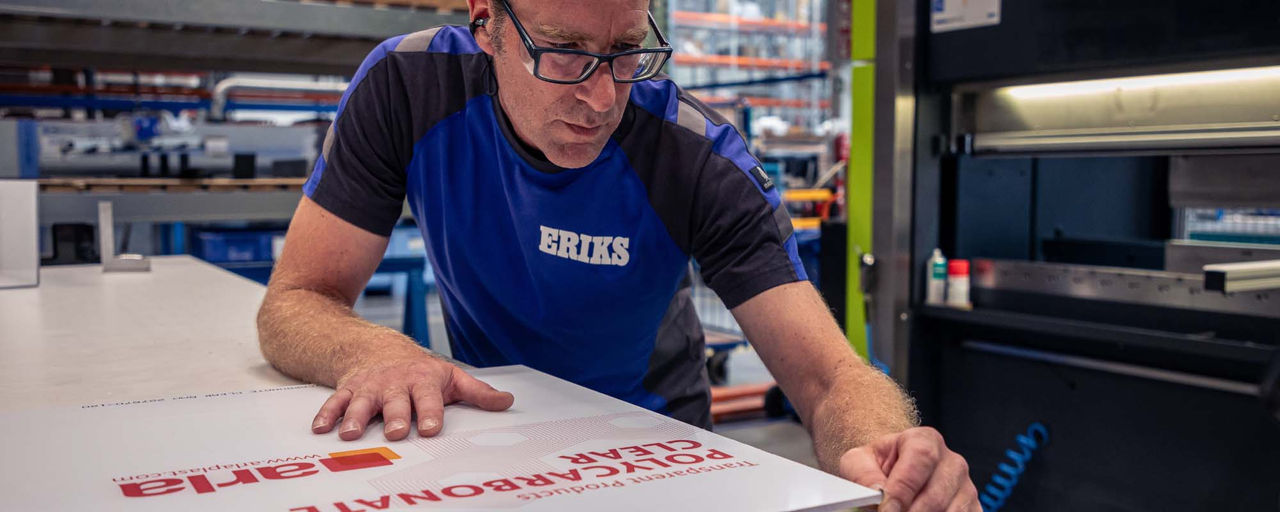 Ein ERIKS Mitarbeiter in blauer Arbeitskleidung prüft sorgfältig eine Polycarbonatplatte auf einer Werkbank in einer Lagerumgebung.
