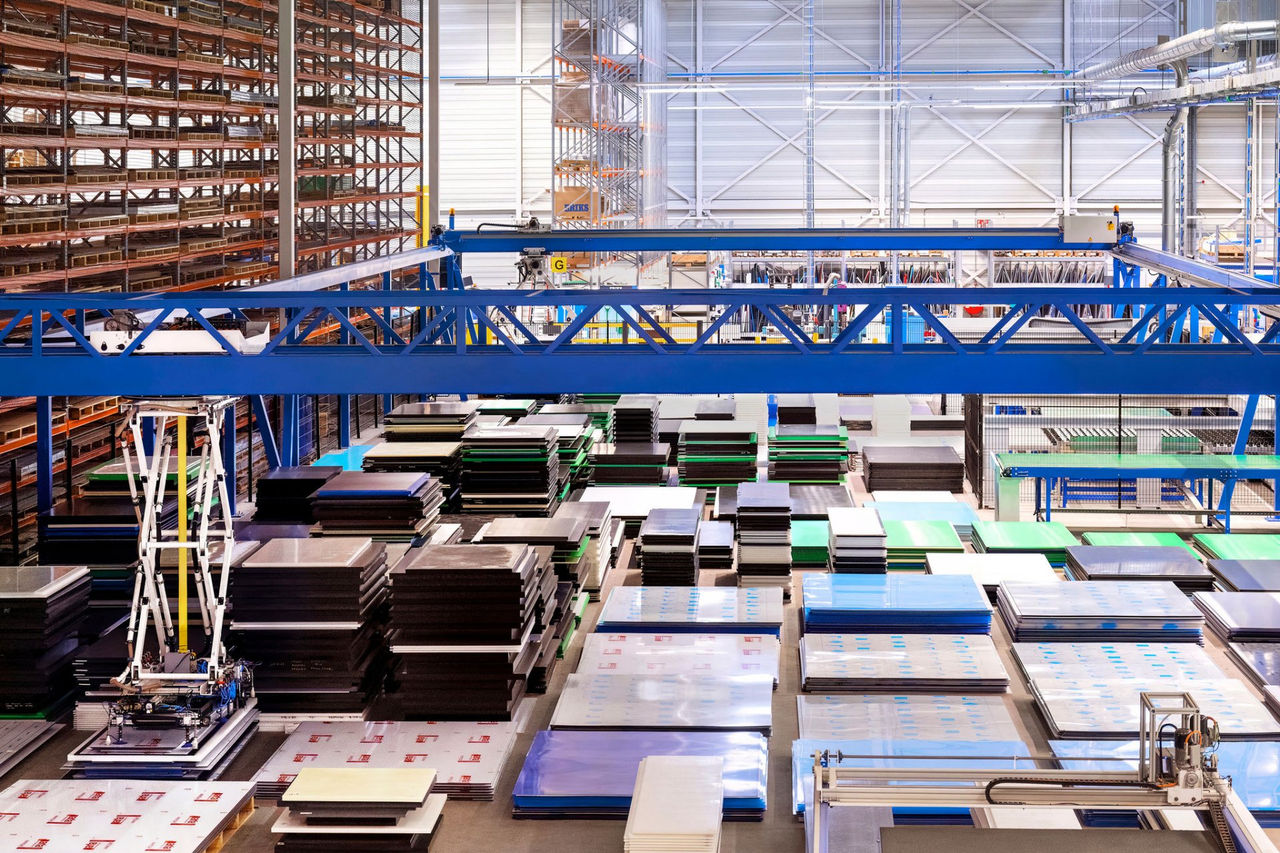 Ein geräumiges Lager, gefüllt mit ordentlich gestapelten Kunststoffplatten in verschiedenen Farben und Größen. Ein blauer Brückenkran ist zu sehen, im Hintergrund stehen Regale und Industrieanlagen.