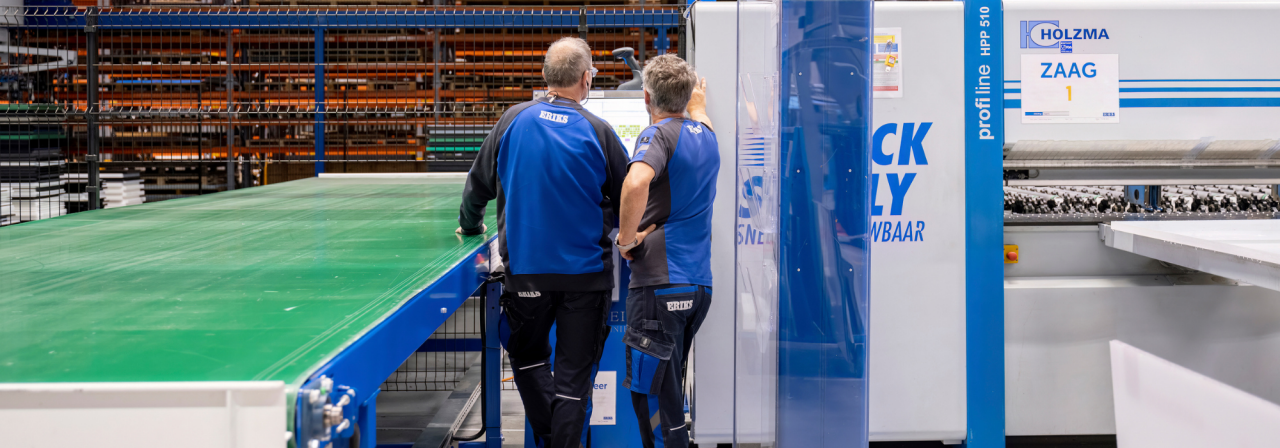 Twee ERIKS-medewerkers in blauwe uniformen bedienen een zaagmachine met het label "ZAAG 1" in een ruime industriële werkruimte met groene werktafels en georganiseerde stellingen op de achtergrond.