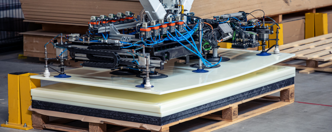 Linkedin Post - Shoes (600 x 600 px) - 4 Een robotisch vacuüm-hefsysteem met meerdere zuignappen en ingewikkelde bedrading, geplaatst bovenop een stapel plastic platen op een houten pallet in een magazijn.
