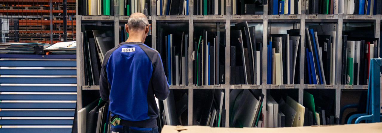 Een ERIKS-medewerker in een blauw uniform staat voor schappen gevuld met diverse plastic platen in verschillende kleuren en maten en organiseert materialen in een magazijn.